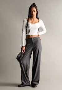 Bílé dlouhé rukávy zkrácený top, šedé pruhované široké kalhoty a černé baleríny. Modelka drží šedou kšiltovku, která ukazuje minimalistický design.