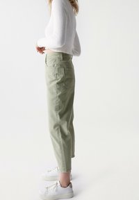 Světle zelené, vysoké tapered kalhoty s předními kapsami, zapínáním na knoflík a jednou zadní kapsou, v kombinaci s bílými teniskami.