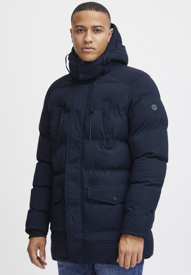 Navy blue puffer jacket with a high collar, drawstring hood, multiple buttoned chest pockets, and a textured finish.