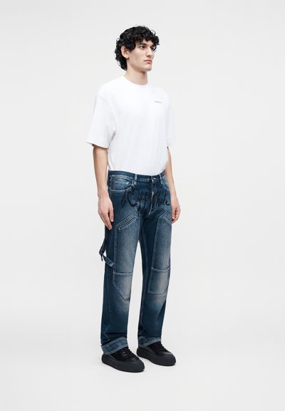 Jeune homme aux cheveux bouclés portant un t-shirt blanc surdimensionné, un jean bleu foncé avec des détails en patch et des baskets noires, debout devant un fond blanc.