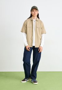 Beige kortermet skjorte med to framover-lommer, lagd over en hvit langermet skjorte; mørk blå løstsittende jeans og brune sneakers med blå snøringer.