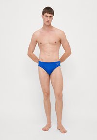 Blå svømmeshorts laget av glat, elastisk materiale, med en slank design og en liten logo på midjebåndet. Enkel og minimalistisk stil.