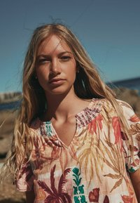 Vaaleahiuksinen nainen, jolla on pitkät hiukset ja kukkakuvioinen pusero, seisoo ulkona kivikkoisen rannikkomaiseman äärellä kirkkaan sinisen taivaan alla.