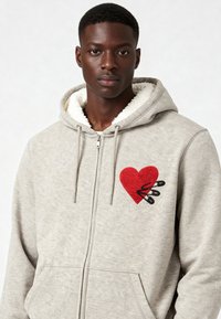 Haut à capuche gris zippé avec une capuche doublée en sherpa, arborant un cœur rouge brodé et des épingles noires sur le côté gauche de la poitrine. Deux poches avant.