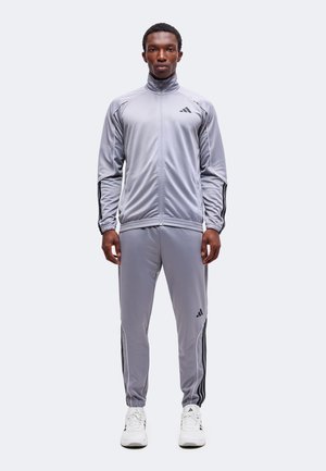 Uomo che indossa una tuta Adidas grigia con strisce nere e scarpe da ginnastica Adidas bianche, in piedi di fronte su uno sfondo semplice.