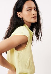 Blouse sans manches de couleur jaune clair en tissu doux, avec un col boutonné et une coupe décontractée, présentant des coutures visibles et une silhouette légèrement surdimensionnée.