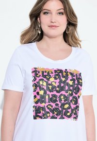 Hvid T-shirt med rund halsudskæring, der har et dristigt grafisk print i sort, pink og guld. Blødt stof, korte ærmer, afslappet pasform.