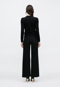 Schwarzes langärmliges Oberteil kombiniert mit weitem schwarzen Jeanshosen, die ein figurbetontes Design und eine strukturierte Stoffqualität aufweisen, ergänzt durch schwarze High-Heel-Schuhe.