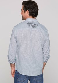Camisa de manga larga azul claro con un patrón texturizado, que presenta un logo en la parte posterior y puños remangados que revelan un interior estampado.