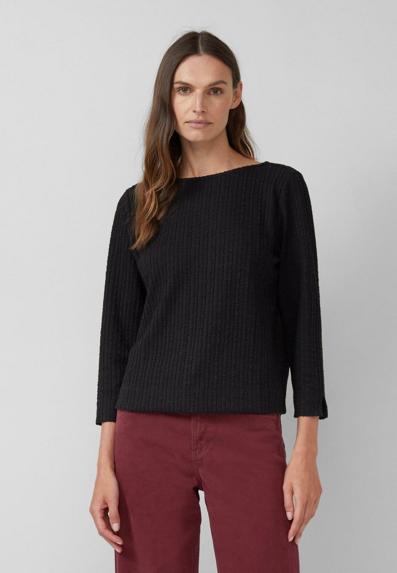 Vrouw met lang bruin haar, gekleed in een zwarte gestructureerde langemouwen top en bordeauxrode broek, staande tegen een effen grijze achtergrond.