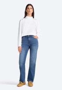 Blouse blanche à manches longues avec col à volants et accents de boutons, associée à un jean en denim bleu taille haute et des chaussures en daim beige.