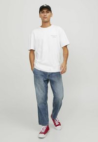 Camiseta blanca de algodón con hombros caídos y logo en el frente, combinada con vaqueros de denim azul claro y zapatillas rojas de caña alta.