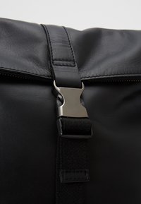 Sac en cuir noir avec fermeture par boucle en métal argenté et détail de sangle tissée noire en gros plan.