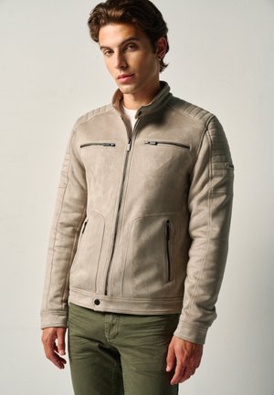 Young man wearing a light beige suede jacket with zippered pockets and olive green pants, standing against a plain light background.