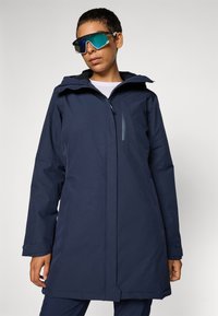 Marinblå vattentät parka med huva, framdragkedja och sidofickor. Slät textur och subtila sömdetaljer, bärs med reflexsolglasögon.