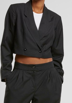 Vrouw draagt een zwarte cropped double-breasted blazer en een zwarte broek met hoge taille, met haar handen in de zakken en een wit topje eronder.