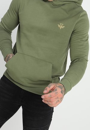 Person trägt olivgrünen Hoodie mit Kängurutasche und kleinem goldbesticktem Emblem, kombiniert mit schwarzen strukturierten Hosen.