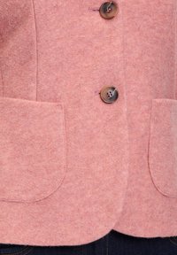 Vaaleanpunainen villablazer, jossa on kaksi suurta pyöreää taskua, kolme pyöreää nappia ja pehmeä tekstuuri. Muotoiltu siluetti, jossa on hieman kaareva helma.