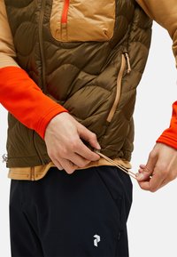 Gesteppteste olivegrüne Weste mit sandfarbenen Akzenten und einer Fronttasche. Modell passt den Kordelzug des cremefarbenen Shirts darunter an. Orangefarbene lange Ärmel sind sichtbar.