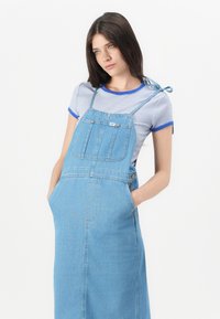 Abito a salopette in denim di colore azzurro chiaro con bretelle regolabili, grande tasca frontale e silhouette dritta. Accompagnato da una camicia a righe.