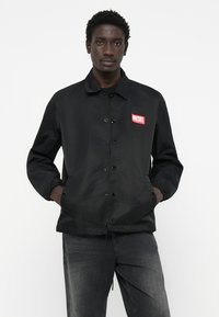 Svart nylonjakke med knappefront, elastiske mansjetter og et rødt "DIESEL" logo-lapp på brystet. Uformell, lett design.