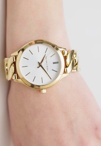 Goudkleurig horloge met een witte wijzerplaat, eenvoudige uurmarkeringen en een chunky schakelarmbanddesign. Voorzien van een kroon voor het instellen van de tijd.