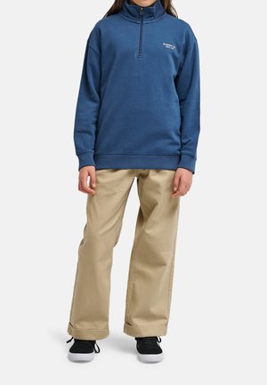 Persoon die een blauwe trui met kwartsluiting draagt, beige broek met wijde pijpen en zwarte sneakers, staand tegen een witte achtergrond.