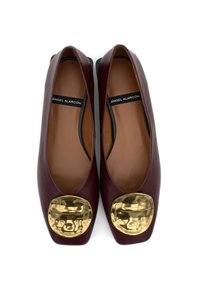 Ballerine in pelle bordeaux con una brillante finitura metallica dorata circolare sulla punta. Texture liscia con un design piatto e arrotondato.