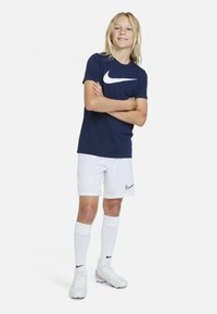 Nike sportovní oblečení: tmavě modré tričko s bílým logem, bílé šortky, bílé fotbalové ponožky a bílé kopačky s černými akcenty.