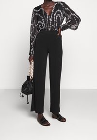 Blusa nera con motivo a fantasia, maniche lunghe, scollatura a V e dettagli floreali, abbinata a pantaloni neri a vita alta e sandali neri.