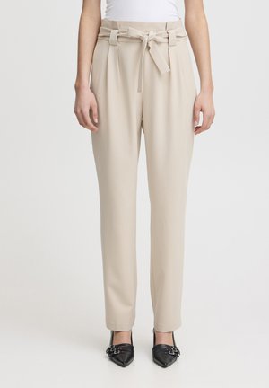Pantaloni beige a vita alta con cintura annodata, tessuto liscio, gambe larghe e pieghe frontali. Abbinati a scarpe nere a punta.