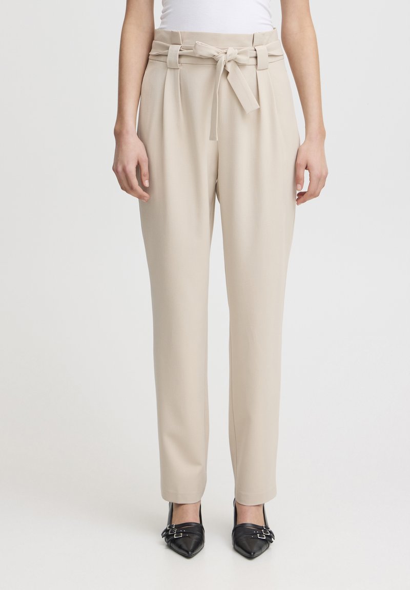 Pantalons beiges taille haute avec une ceinture nouée, tissu lisse, jambes larges et plis sur le devant. Associés à des chaussures noires à bout pointu.