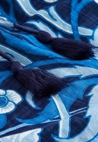 Tessuto blu con motivo floreale, caratterizzato da nappe blu scuro su ciascuna estremità. La trama è morbida, con diverse tonalità di blu e dettagli bianchi.