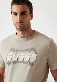 Beige puuvillane T-särk, mille esiküljel on suur, uduseks muutunud valge "GUESS" logo. Ümar kaelus ja lühikesed varrukad. Sile tekstuur.