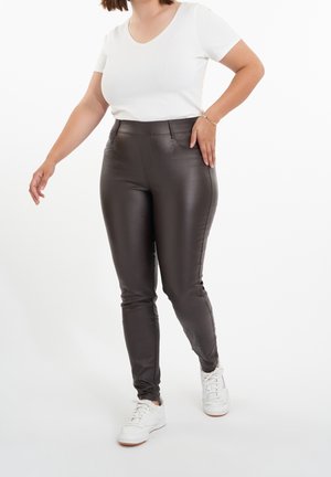 Femme portant un pantalon en cuir marron foncé, un t-shirt blanc à manches courtes, des baskets blanches et un bracelet doré sur fond blanc.