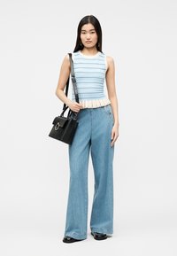 Frau trägt ein ärmelloses blau-creme gestreiftes Oberteil, hoch taillierte, weit geschnittene Jeans, schwarze Schuhe und trägt eine schwarze Handtasche über der Schulter.