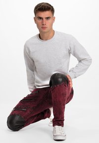 Sweatshirt gris et jean bordeaux avec des accents en cuir noir aux genoux, des zippers et des détails usés. Le modèle est à genoux.