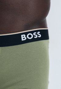 Grüne Boxer-Shorts mit einem schwarzen Bund, der das Logo "BOSS" in Weiß und goldenen Akzenten trägt. Das Material sieht glatt und dehnbar aus.