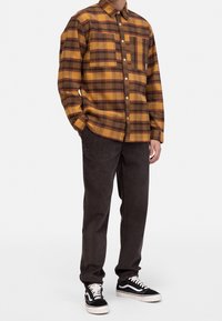 Hombre con camisa de cuadros amarilla y marrón, pantalones marrón oscuro y zapatillas negras y blancas, de pie con una mano en el bolsillo.