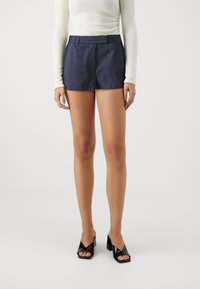 Pantalones cortos marinos a rayas con un ajuste entallado y cinturilla, combinados con zapatos de tacón de charol negros, sobre un fondo neutro.