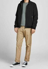 Veste noire à fermeture éclair, T-shirt gris, chinos beige et baskets noires avec semelles blanches. La tenue présente un design simple et moderne.