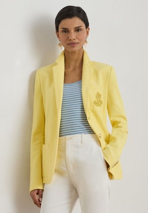 Femeie purtând un sacou galben peste un top cu dungi albastre și albe, cu pantaloni albi, stând în fața unui perete deschis la culoare, simplu.