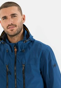 Blaue wasserfeste Jacke mit Kapuze, Frontreißverschluss und zwei Reißverschlusstaschen. Mit Zugbändern und kontrastierender Innenauskleidung.