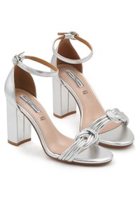 Sandali argentati con tacco, presentano una striscia anteriore annodata, una cavigliera con fibbia e un tacco quadrato spesso. Realizzati in materiale sintetico liscio.