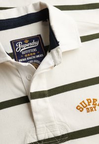 Chemise à col rayée blanc et vert olive avec broderie jaune "Superdry" et étiquette bleu marine à l'intérieur du col portant l'inscription "Superdry Outfitters".