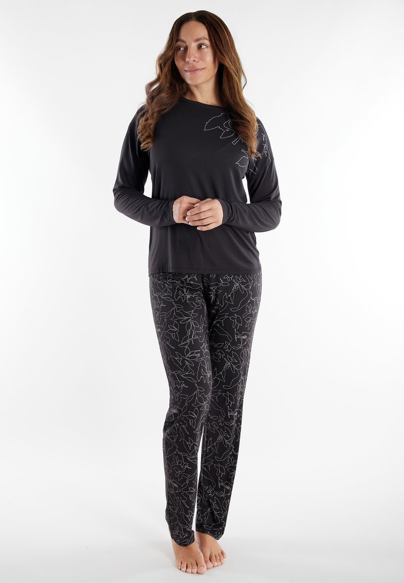 Woman standing barefoot wearing black long-sleeve top and black pants with white abstract leaf pattern against white background.