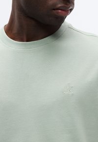 Κοντινό πλάνο ενός άνδρα που φοράει ένα ανοιχτοπράσινο μπλουζάκι με στρογγυλή λαιμόκοψη και με μικρό λευκό λογότυπο "ck" κεντημένο στο στήθος.