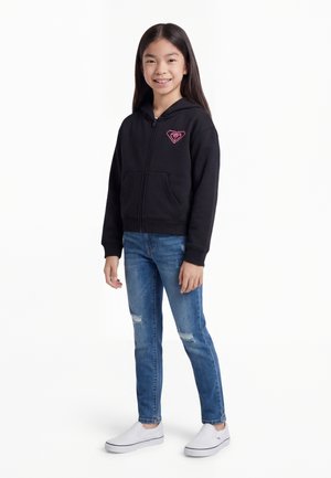 Dívka stojí a usmívá se, má na sobě černou mikinu na zip s růžovým logem, roztrhané modré džíny a bílé nazouvací boty na bílém pozadí.