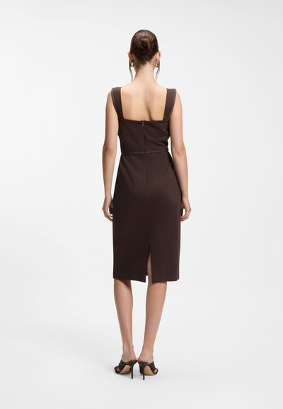 Femme avec les cheveux en chignon portant une robe midi sans manches marron foncé avec une fermeture éclair au dos et une fente, associée à des sandales noires à talons hauts.