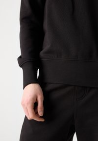 Černá mikina a kalhoty s žebrovanými manžetami a lemem, vyrobené z texturované látky. Detail ruky opírající se o pas, který ukazuje detaily oděvu.